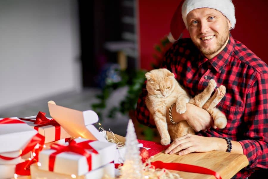 Man in a Santa hat holding an orange cat while sitting at a table with wrapped Christmas gifts.