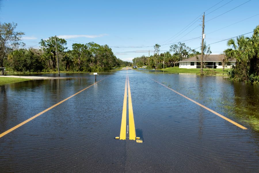 Flooded street with water covering the roadway near houses and trees.
